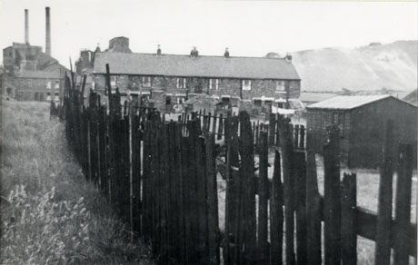 Photograph showing in the foreground the fence of an allotment, running away from the camera; in the distance is a row of terraced houses and, behind it on its left, a pit heap and, behind it on its on its right, a large building and two tall chimneys; the photograph has been identified as Shotton Colliery