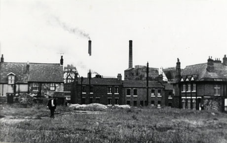 Photograph showing an area of open ground covered in rough grass, across which a man is walking; behind the open area are a row of houses and, at the side, a large house, possibly a public house; behind the row of houses are the winding gear and two chimneys of a colliery, partly obscured by smoke or steam; the colliery has been identified as Shotton Colliery