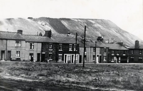 Photograph of an open area of rough grass with a terrace of houses behind it , running across the picture; behind the houses the side of a pit heap, overshadowing the houses, can be seen; the photograph has been identified as Shotton Colliery