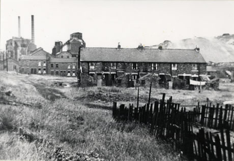 Photograph showing the same view as shot0027, but showing more of the buildings of the colliery, as the photograph is taken further to the left; a small portion of the fence is on the right, but more of the rough ground to the side of the terrace of houses can be seen; beyond the rough ground two more terraces and the large buildings and two chimneys of the colliery are in view; the colliery has been identified as Shotton Colliery
