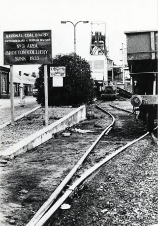Photograph showing on the extreme left a line of low buildings, in front of which is a road and in front of the road a sign reading: National Coal Board Northumberland and Durham Division No 3 Area Shotton Colliery Sunk 1833; beyond the sign are colliery buildings and to the side of it a rail track on which a small steam locomotive is standing facing the camera in the middle distance; the right of the picture is occupied by part of the rear of a coal waggon; the steam locomotive has been described as being out of use at the time of the photograph