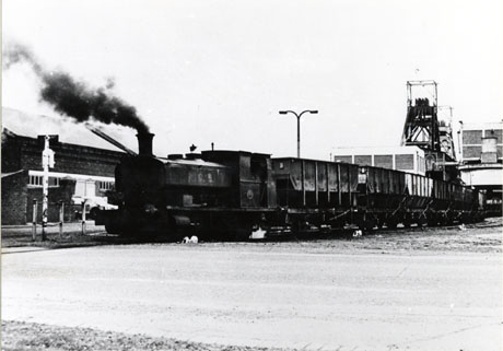Photograph showing a steam locomotive pulling a line of coal waggons from right to left of the picture; behind the waggons are colliery buildings at Shotton Colliery