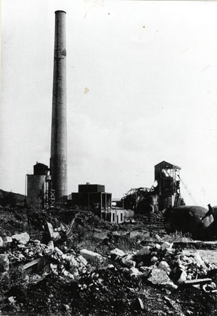 Photograph showing rubble-strewn ground in the foreground and partly demolished colliery buildings in the middle distance; in the middle of the buildings is a tall chimney standing out against the sky; it has been identified as Shotton Colliery Pit Chimney Just Before Demolition