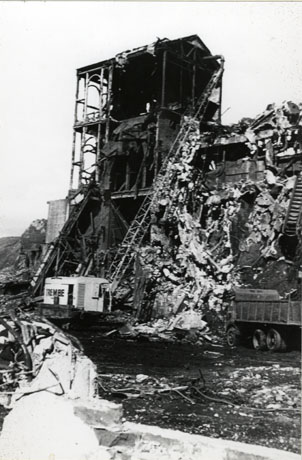Photograph showing debris in the foreground and a large high structure in the middle distance with only its skeleton intact; piles of debris are at its side; a mobile crane and a lorry are in front of the building and debris; the picture is identified as Pulling Down The Coke Ovens, Shotton Colliery