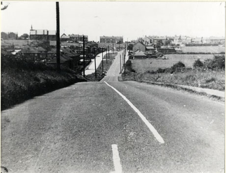 Photograph showing a tarmacadamed road running downhill away from the camera towards a group of houses on the horizon; an estate of semi-detached houses runs along the left of the road and there is a church on the left on the horizon; the road has ben identified as Road into Haswell from Shotton