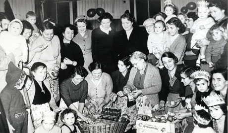 Photograph of approximately twenty women dressed in frocks, hats and aprons, standing round eight women who are sitting in a semi-circle with six children, wearing party hats, a box of apples and a basket in which there are presents; the wall, which appears to be constructed of wood or corrugated iron, has balloons on it; the party is described as The Last Child Welfare Party Held In The Miners' Institute, Shotton Colliery