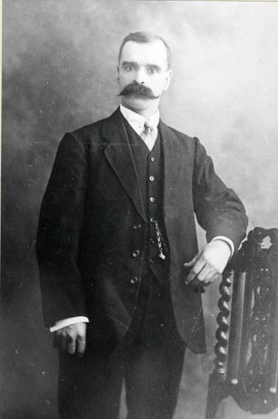 Photograph of a man with a walrus moustache wearing a suit, waistcoat, collar and tie and watch chain, with his left arm resting on the back of a wooden chair; he has been identified as Bill Henderson of Shotton Colliery