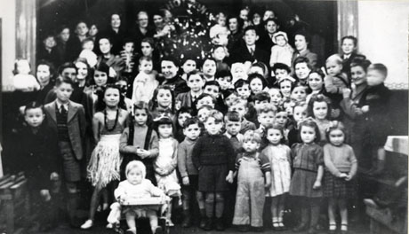 Photograph of approximately twenty women and approximately forty children grouped on a stage and in front of the stage in a hall; one of the small girls is dressed as a South Sea girl in a grass skirt; in the middle of the group there is an indistinct shiny object, possibly a Christmas tree; the event has been identified as Shotton Colliery Mothers' Club Children's Party