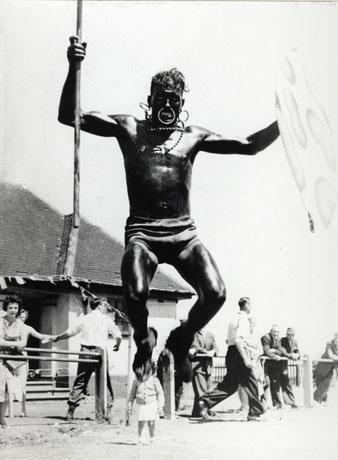 Photograph of a man leaping into the air wearing only briefs and brandishing a pole in his right hand; he has blacked his skin and has rings in his nose and ears; behind the man is part of the roof and walls of a low building and six men, two women and a child; the event has ben identified as Shotton Colliery's First Annual Children's Sports Day, 1955