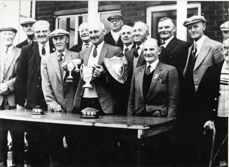 Photograph showing thirteen elderly men standing behind a table with a wall with windows behind them; one man is holding three trophy cups and another man is holding a shield; two rosebowls are on the table; the men are dressed in jacket, collar and tie; they have been identified as Shotton Colliery Veterans at a Bowls Club Presentation
