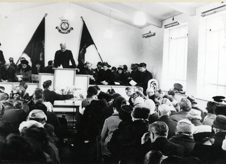 Photograph of the interior of a hall with windows down the right hand side; at the end of the room facing the camera is a dais on which there are a man in the centre and approximately twelve people sitting either side of him; rows of people are sitting in the hall facing the dais and away from the camera; behind the man on the dais on the wall are two crossed flags and the badge of the Salvation Army; the event has been identified as A Salvation Army Service, Shotton Colliery