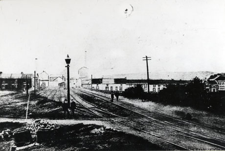 Photograph showing railway lines running away from the camera, with two rows of terraced houses at right angles to the rails on either side; a very indistinct picture of a tall chimney and colliery building can be seen beyond the houses; a couple of indistinct figures can be seen on either side of the rails; the scene has been identified as Shotton Colliery 1902