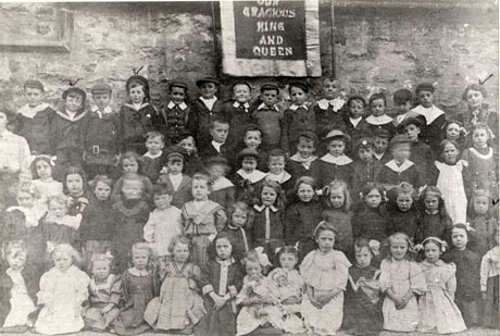 Photograph of approximately sixty children, between the ages of three and seven, posed in front of a rough stone wall with a banner on it reading Our Gracious King and Queen; two women are standing either side of the group; the children have been identified as Sunday School Children at St. Saviour's Church Hall, Shotton Colliery, about 1910; it is possible that the banner indicates that the children were marking the Coronation of King George V in 1910; the boys, second and fourth from the left on the back row, have been identified as Dr. Gross's sons, and the woman on the right of the group as Edith Millington