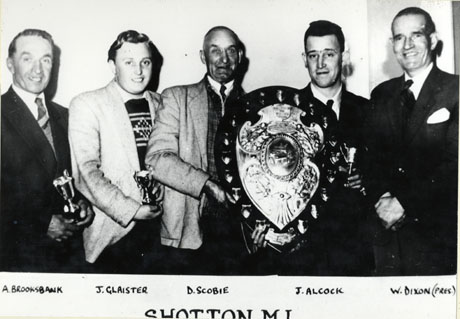 Photograph of five men wearing jackets and ties, posed in a row; one is holding a large shield and two are holding small trophy cups; they have been identified as, from right to left: A Brooksbank; J. Glaister; D. Scobie; J. Alcock; W. Dixon (President); the occasion has been identified as The Northern Amateur Billiards League Presentation, Shotton Miners' Institute, 1958-9