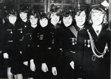 Photograph of eight girls, aged approximately fourteen years, in uniform, posed in a line in front of a panelled wall in an unidentified building; they wear medals, chevrons denoting status, and pointed hats; the girl at the front of the row is wearing an elaborate hat and an elaborate lanyard and is presumably the leader of the group; they have been identified as members of Shotton Colliery Girls' Brigade, 1950s