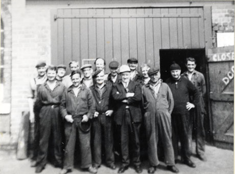 Photograph of fourteen men in overalls standing outside the door of a workshop; one of them is wearing a hard hat and the rest caps; they have been identified as Shotton Colliery Group
