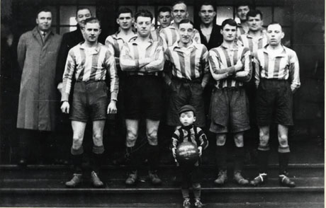 Photograph of eleven members of a football team standing on some steps, accompanied by two men and a small boy, standing in front of the group, holding a football; they have been identified as follows: Back Row, left to right: Mr. Summerson; Mr. Jackson; Mr. Hamilton; Mr. Wrightson; Mr. Connelly; Front Row, left to right: Mr. Levinson; Mr. Tucker; Mr. Knaggs; Mr. Peel; the boy: Peter Nord; the picture has been described as Shotton Colliery Charity Football Team, Christmas, early 1930s