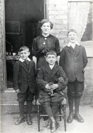 Photograph of a woman standing in front of the open door of a house with two boys, aged approximately twelve and nine years, standing either side of her; a boy, aged approximately ten years, is sitting on a chair in front of her holding a small dog, which appears to be determined to wriggle away; a table and cups can be seen through the doorway; they have been identified as Mr. Arkwright (right) with his sister and brothers in George Street, Shotton Colliery