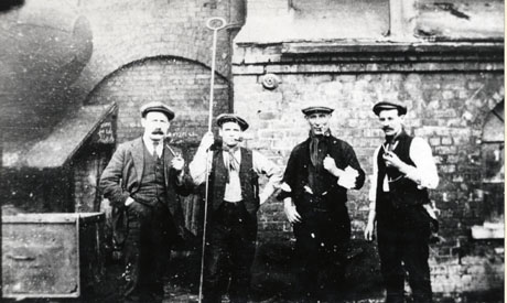 Photograph of four men standing in front of the wall of a large brick building; three are wearing working clothes and the fourth a suit; all four are smoking pipes and one is holding a long metal pole with a circle at the end; they have been identified as Shotton Colliery Officials, Firing Boilers During The 1921 Strike