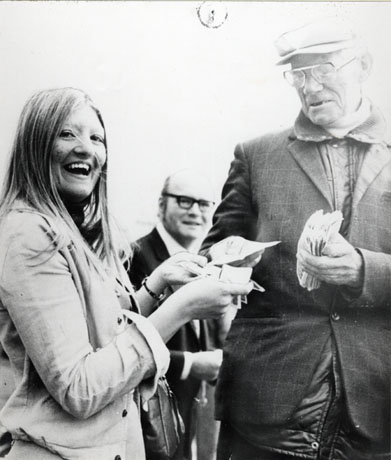 Photograph showing a young girl at the left of the photograph laughing and handing papers or banknotes to a middle-aged man on the right of the photograph who is wearing a jacket on top of a padded jacket, and holding a wad of bank notes; the head and shoulders of another man can be seen in the middle of the photograph behind the other two; the photograph has been described as Johnny Ridley, Bookmaker of Shotton Colliery, at York Races