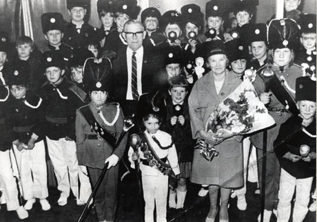 Photograph of approximately thirty children in a uniform of light trousers, dark tunic, and busbies, posed in front of a wall; in front of the group are three children wearing sashes over their uniforms and carrying maces; also in front of the thirty children are a middle-aged man and a middle-aged woman who is carrying a bouquet of flowers; they have been described as Shotton Colliery Jazz Band