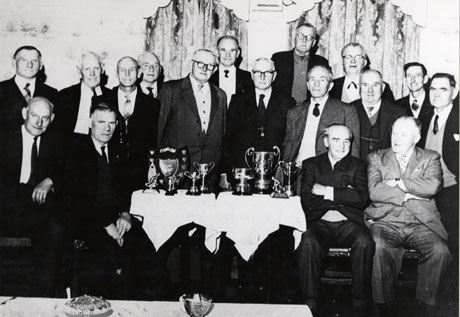 Photograph of thirteen elderly men standing behind a table on which ten trophies are displayed; two elderly men are sitting on either side of the table; at the front of the picture there is a table top on which there are a sugar bowl and another unidentifiable item; the photograph has been described as Bowls Club Presentation, Shotton Colliery