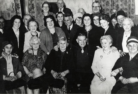 Photograph of a group of eight elderly men and seventeen elderly women posed in front of an indoor wall with windows and curtains; they are all dressed in suits and smart dresses; the photograph is described as Women's Institute, Shotton Colliery
