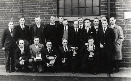 Photograph of a group of eighteen men, wearing suits, posed outside a brick building; five of the men are kneeling in front of the rest and are holding trophies: two large cups, two small cups, a rose bowl and a canteen of knives; the men have been identified as members of Shotton Comrades Homing Pigeon Club