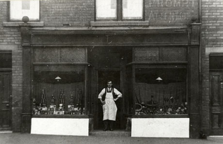 Photograph of a shop front with two windows, but with no name above the shop front; tinned goods can be seen piled in the left hand window and fruit in the right hand window; a man wearing a white apron is standing in the doorway; the shop has been described as Mr. Fawell's Shop, Front Street; the man has not been identified
