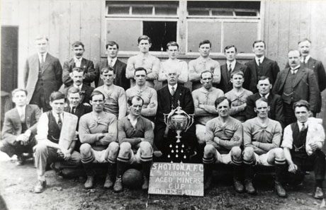 Photograph of eleven men in football strip accompanied by sixteen other men in suits, two of whom have towels over their shoulders and are, presumably, trainers; in the middle of the group is a large trophy cup, ten medals, and a notice reading Shotton Athletic Football Club Aged Miners Cup Winners 1921-22