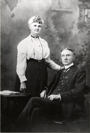 Photograph of a middle-aged woman standing behind a table with her left hand resting on the shoulder of a middle-aged man who is sitting in a chair at the right of the table; the woman is wearing along dark skirt. light coloured blouse and long necklace; the man is wearing a three-piece suit with a high collar; the photograph has been taken in a photographer's studio and depicts Mr. and Mrs. Davison, First Caretakers of Shotton Miners' Institute