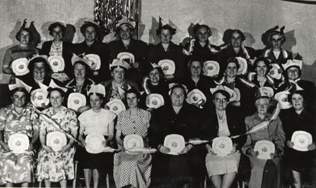 Photograph of twenty three women dressed in summer frocks and paper hats, formally posed in front of an interior wall; they are each holding a commemorative plate and tricolour streamers are lying over the members of the group; they have been identified as members of the Womens' Section of Shotton Colliery British Legion celebrating the Coronation of Queen Elizabeth II