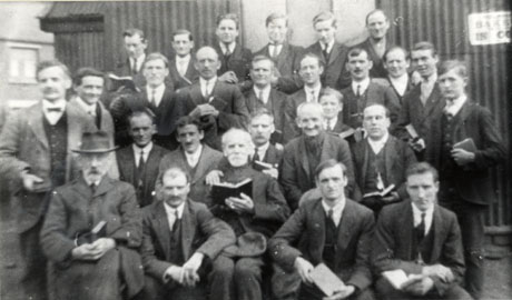 Photograph of twenty seven men posed outside a corrugated iron building; the men range in age from very young to very old and all are carrying a book; they are all dressed formally in suits and may be Lay Preachers, as they have been identified as members of the Wesleyan Methodist Church, Shotton