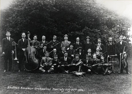 Postcard photograph, of eighteen men, one woman, and five boys, posed on grass in front of trees; most of the members of the group are carrying musical instruments; the photograph is entitled Shotton Amateur Orchestral Society, July 20th 1924