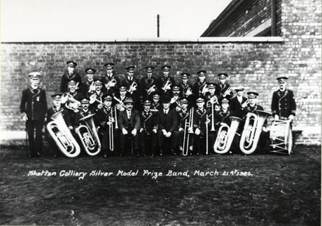 Photograph of twenty one men and three boys in uniform, carrying musical instruments, posed against an exterior brick wall; they are accompanied by two men in ordinary clothes; the photograph is inscribed Shotton Colliery Silver Model Prize Band, March 21st 1926