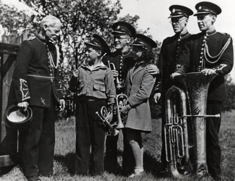 Photograph showing an elderly man on the left of the photograph dressed in a uniform and carrying a peaked cap; he is talking to a boy and a girl, aged approximately eight and nine, who are wearing peaked caps and carrying trumpets; behind the children is a man wearing a uniform, with a hand on the shoulders of each of the children; on the right of the photograph are two men dressed in uniform watching the other figures; one of these men is resting a large brass instrument on the ground; the photograph is described as depicting members of the Hearn family, members of Shotton Colliery Silver Prize band