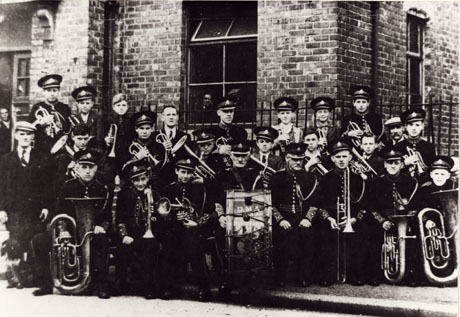 Photograph of seventeen boys and eight men in uniform and carrying instruments, posed outside a brick building with railings immediately behind them; people can be seen looking out of the windows and doorway of the building, which has been identified as Shotton Miners' Institute; the band has been identified as the Prize Silver Band