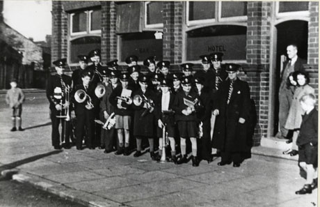 Photograph of approximately fifteen boys wearing peaked caps and carrying musical instruments, accompanied by six men in uniform, also carrying musical instruments; the group is standing outside a public house, identified as the Station Hotel, Shotton; three children and a man in the doorway of the public house are watching the group; the group is described as Shotton Colliery Band, but its members seem to be wearing the same uniforms as the members of the band in shot0077, 78, 79,; the photograph has been identified as having been taken on Armistice Day 1942, an event unknown to history, so it may be that the occasion for the photograph was Victory in Europe Day, 8 May 1945; the hypothesis that the photograph was taken during the era of the Second World War is borne out by the fact that most of the members of the band are children