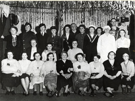 Photograph of twenty two women and one man, posed on a stage with tinsel and a partly-furled flag behind them; the women on the back row are in uniforms of the various armed services and nursing; the women on the two rows in front are wearing blouses and skirts; the man on the back row is holding an accordion; the group has been identified as a British Legion Concert Party at Shotton Colliery with Mrs Winning on the left of the front row
