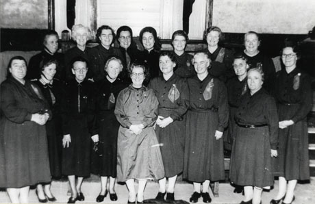 Photograph of eighteen middle-aged women posed in front of a stage; six of the women are wearing Salvation Army uniform; the rest are wearing dresses with belts and cloths pinned to their left shoulders; they are described as Shotton Colliery Salvation Army Home League Singers