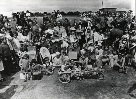 Photograph of children in fancy dress as part of a carnival at Shotton Colliery; the costumes include Catweazle; Dairy Lea Cheese Spread; native Americans; Micky Mouse; a Mexican; a bride and other characters impossible to determine; the children are being watched by other children and adults; the people are in front of a field