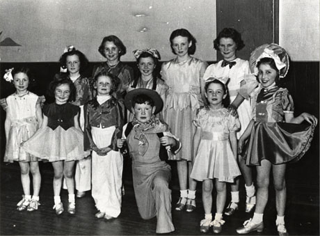 Photograph of ten small girls and one small boy standing in a group inside a hall; the girls, who are aged approximately six years to fourteen years, are dressed in elaborate party frocks; the boy, aged approximately seven years, appears to be dressed as a cowboy; they have been described as Mrs. Winning's Children's Concert Party in a Comic Pantomime at Shotton Colliery