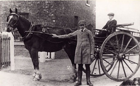 Photograph showing a horse in the shafts of a two-wheeled vehicle, identified as a trap; a man is standing in front of the horse which is seen from the side; he is holding one of the shafts and is wearing a cap, long jacket, and gaiters; a boy is sitting in the driving seat of the trap; behind the horse and trap, the corner of a brick building can be seen; it has been identified as White House Farm; the pony and trap is described as Hutchinsons' Pony and Trap