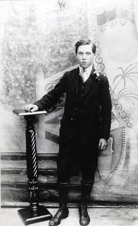 Photograph, taken in a photographer's studio, of a boy of approximately sixteen years wearing boots, gaiters, a jacket with a button hole and a tie; he is posed against a painted cloth and is resting his right hand on a pedestal; he hs been identified as John R. Hutchinson of Shotton Colliery