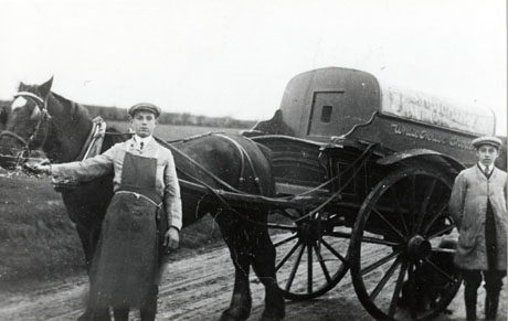Photograph of a horse in the shafts of a covered cart with two wheels, photographed on a country road with a field behind; the words: S. Hutchinson White House Shotton can be seen on the cart; a boy, aged approximately sixteen years wearing an overall and an apron, is holding the horse's head; another boy, also aged approximately sixteen years wearing an overall and gaiters, is standing near the cart; the boy at the horse's head may be the boy depicted in shot0090; the cart has been identified as Hutchinson's Butcher Cart