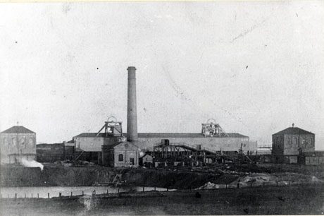 Photograph of the buildings of Shotton Colliery showing a strip of water with the buildings behind it; the buildings include a winding house at either side of the picture, a long low building in the middle with a tall chimney and winding gear