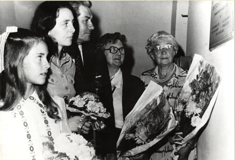 Photograph of a young girl, Julie;a young woman, Miss Proudlock; a man, Billy Lawson; a middle-aged woman, Mrs. Cudlip;and an elderly woman, Mrs. Fairclough; looking at a plaque on a wall; the young woman is holding a posy, the other women are holding large bouquets; the inscription on the plaque cannot be read, but the photograph has been identified as recording the opening of Shotton Community Centre