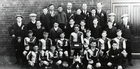Photograph of eleven boys in football strip, accompanied by eleven other boys and by eight men, posed outside a brick building; a large trophy cup is in front of the group and a boy in the middle is holding a board on which fifteen medals are pinned; a football at the front reads: Shotton 1919-1920; the team has been identified as that of Shotton Colliery Boys' School