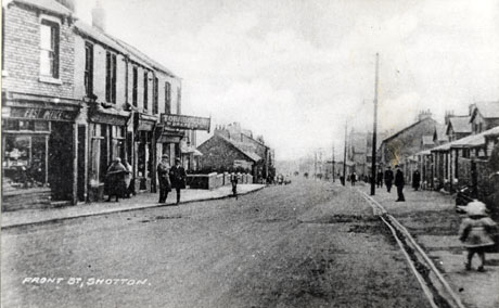 Postcard photograph, entitled Front Street, Shotton, showing a road running downhill away from the camera; on the left side of the road shops, including a Post Office and Turnbull, Drapers, can be seen at the top of the street; further down the sides of small houses can be seen; on the other side of the road, the front of small houses can be seen with large buildings further down the road; indistinct figures can be seen