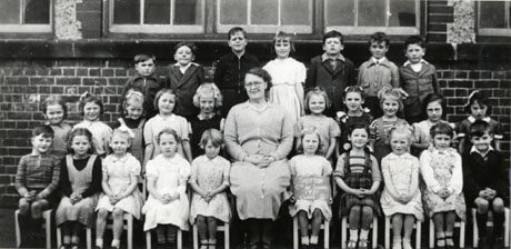 Photograph of a group of twenty six children, aged approximately six years, posed outside a brick building with a woman, presumably their teacher; a child on the front row is holding a notice reading: Shotton County Infants' School 1951 Class 2